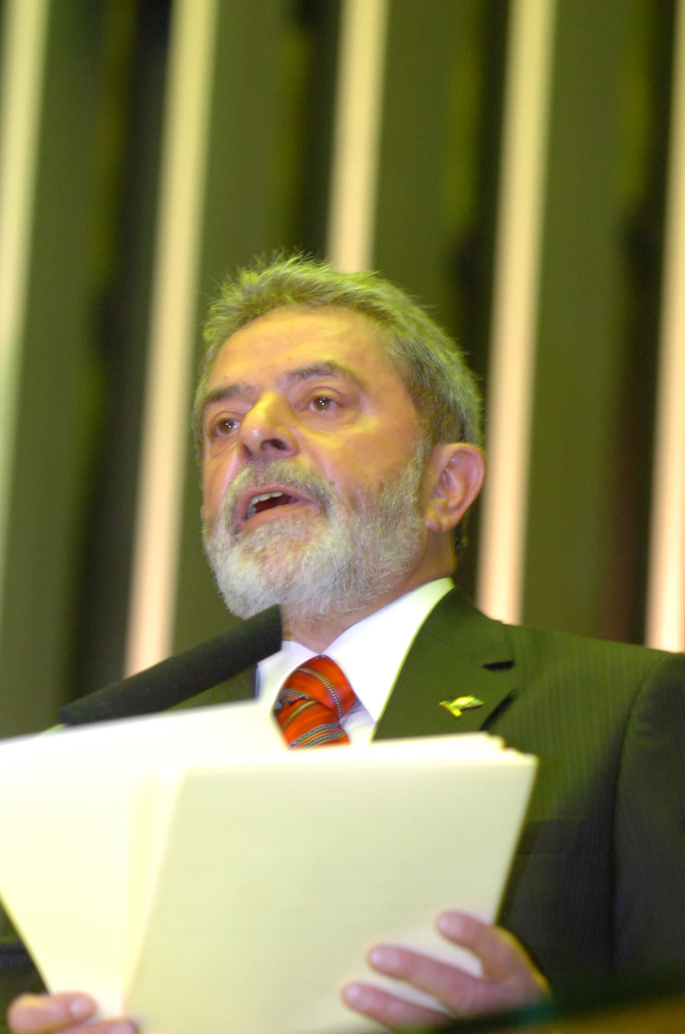 Um homem de barba grisalha, usando terno e gravata, segura folhas de papel diante de um microfone.